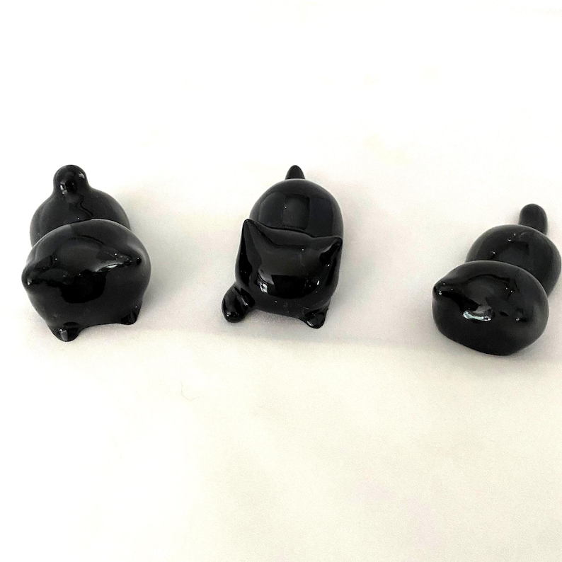 May include: Several glossy black cat ornaments in different poses. One cat is standing, another is seated, and the others are lying down. The ornaments have a smooth, reflective surface and are set against a white background.