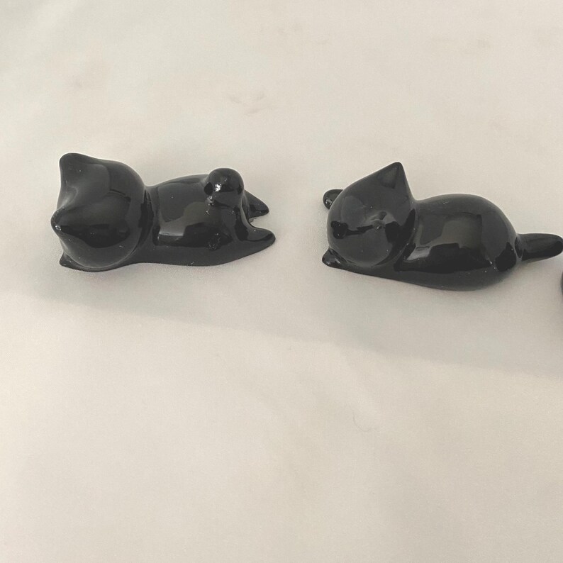 May include: Two glossy black cat ornaments in a relaxed pose. The cats are lying down, with their paws tucked in and tails extended. The ornaments have a smooth, reflective surface, and are set against a plain white background.