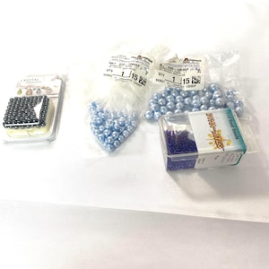 May include: A collection of crafting beads including a small rectangular container of dark gray beads labeled Crystal Passions, two plastic bags filled with light blue round beads, and a clear box of dark blue 11/0 Dyna-Mites seed beads.