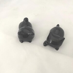 May include: Three small, glossy black ceramic cat figurines. Two are upright, and one is lying on its side. Each cat has a rounded body, small legs, and a tail or ear detail. The figurines are on a white surface.