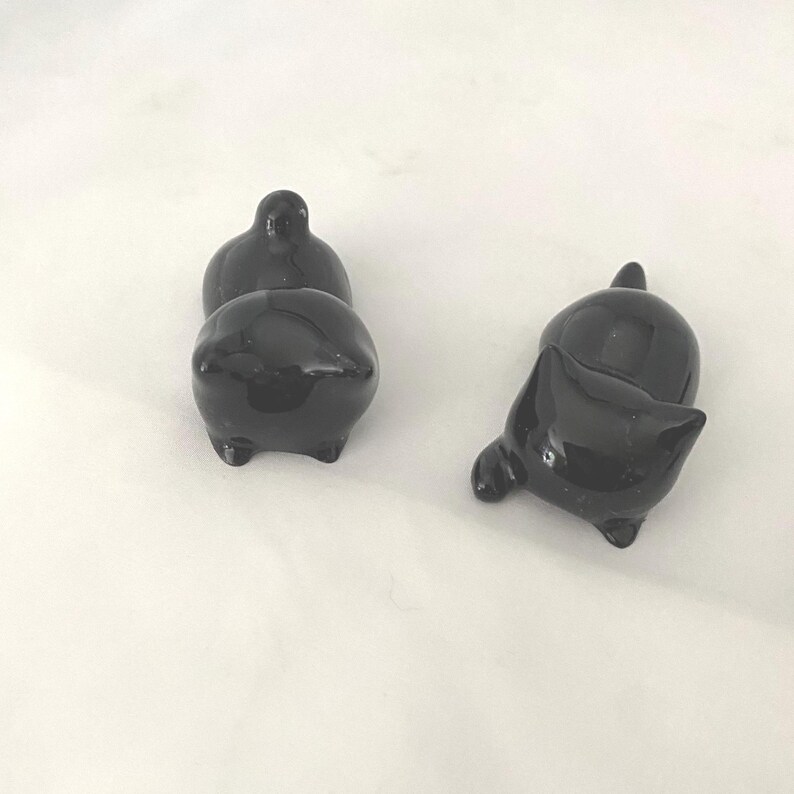 May include: Three small, glossy black ceramic cat figurines. Two are upright, and one is lying on its side. Each cat has a rounded body, small legs, and a tail or ear detail. The figurines are on a white surface.