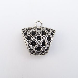 May include: A silver-toned pendant with a geometric design. The pendant features a series of circular black stones set within a diamond-shaped pattern. A small loop is at the top for attaching to a chain or cord.