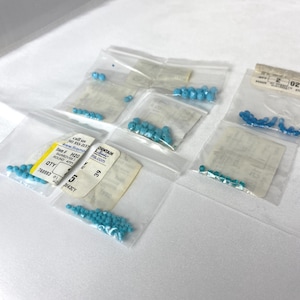 May include: Multiple small clear plastic bags containing turquoise blue round beads. Each bag features a white paper label with printed text including item numbers, quantity, and website information. The beads are uniform in size and color.