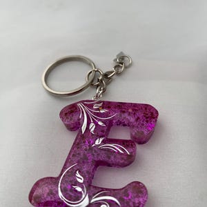 May include: A purple letter "E" keychain with a silver key ring. The letter is made of a translucent material with glitter and silver floral designs. The keychain is a decorative accessory.