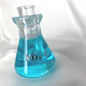 May include: A clear glass flask filled with bright blue liquid. The flask has a clear stopper and the text "NO. 7" is printed on the front. The liquid has a swirling effect, and the flask is set against a white background.