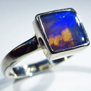 May include: A silver ring with a square-shaped opal gemstone. The opal has a blue and orange iridescent sheen.