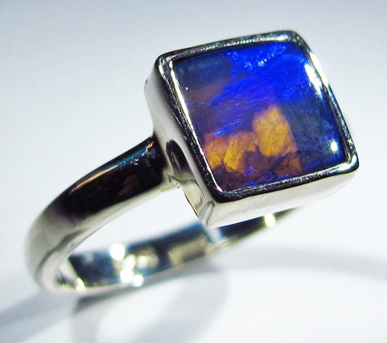 May include: A silver ring with a square-shaped opal gemstone. The opal has a blue and orange iridescent sheen.