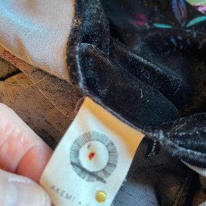 Puede incluir: Una etiqueta blanca con texto negro que dice "AKEMI + KIN" y un logotipo circular con un punto rojo en el centro. La etiqueta est&aacute; unida a una tela de terciopelo negro.