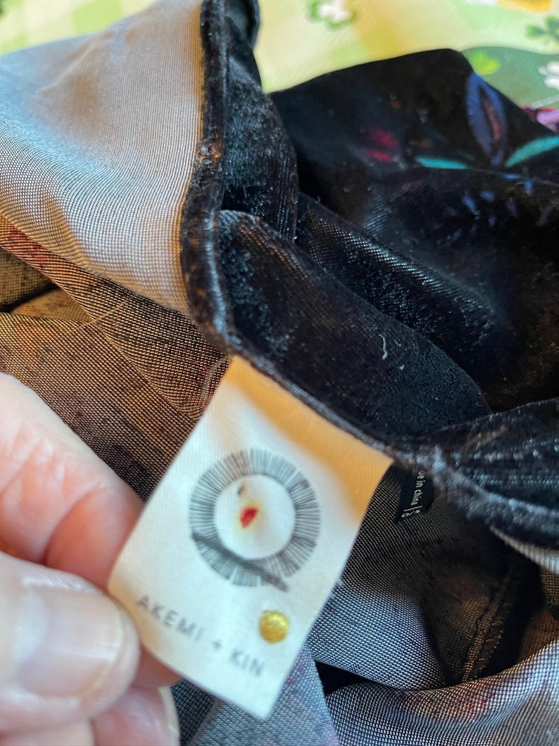 Puede incluir: Una etiqueta blanca con texto negro que dice "AKEMI + KIN" y un logotipo circular con un punto rojo en el centro. La etiqueta est&aacute; unida a una tela de terciopelo negro.