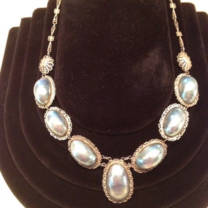May include: A silver-toned necklace featuring large, oval, iridescent blue-gray stones. The stones are set in ornate, filigree-style frames and connected by delicate chain links. The necklace is displayed on a black velvet bust.