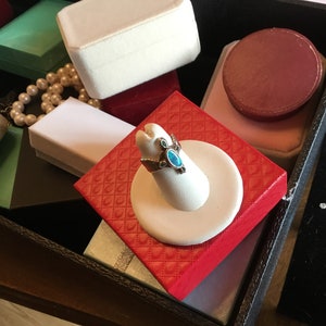 May include: A turquoise ring with a silver turtle design sits on a white ring display stand. The stand is on top of a red velvet box with a quilted pattern. The box is surrounded by other jewelry boxes.