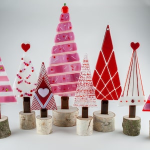 May include: A set of eight glass Christmas trees in various pink, red, and white designs. The trees are decorated with hearts, stripes, and dots. They are displayed on wooden bases.