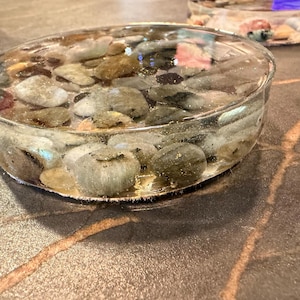 May include: A clear, round resin dish filled with various sizes and colors of smooth stones. The stones are visible through the transparent resin, creating a decorative and natural aesthetic. The dish sits on a textured surface.