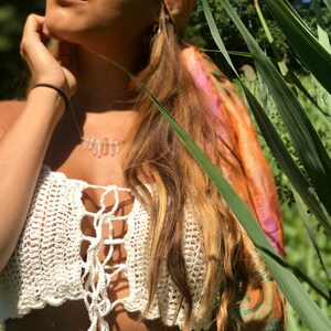 May include: A woman wearing a white crochet top with a tie at the front. She is wearing a brown and orange patterned headscarf and has long blonde hair. The background is a blurred image of green foliage.