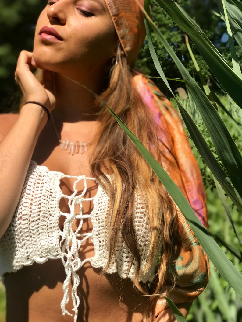 May include: A woman wearing a white crochet top with a tie at the front. She is wearing a brown and orange patterned headscarf and has long blonde hair. The background is a blurred image of green foliage.