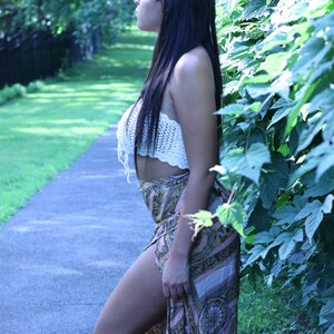 May include: A woman wearing a white crocheted top and a long, patterned skirt. She is standing in a forest setting with green foliage behind her. She is wearing a gold headpiece with small, dangling charms.