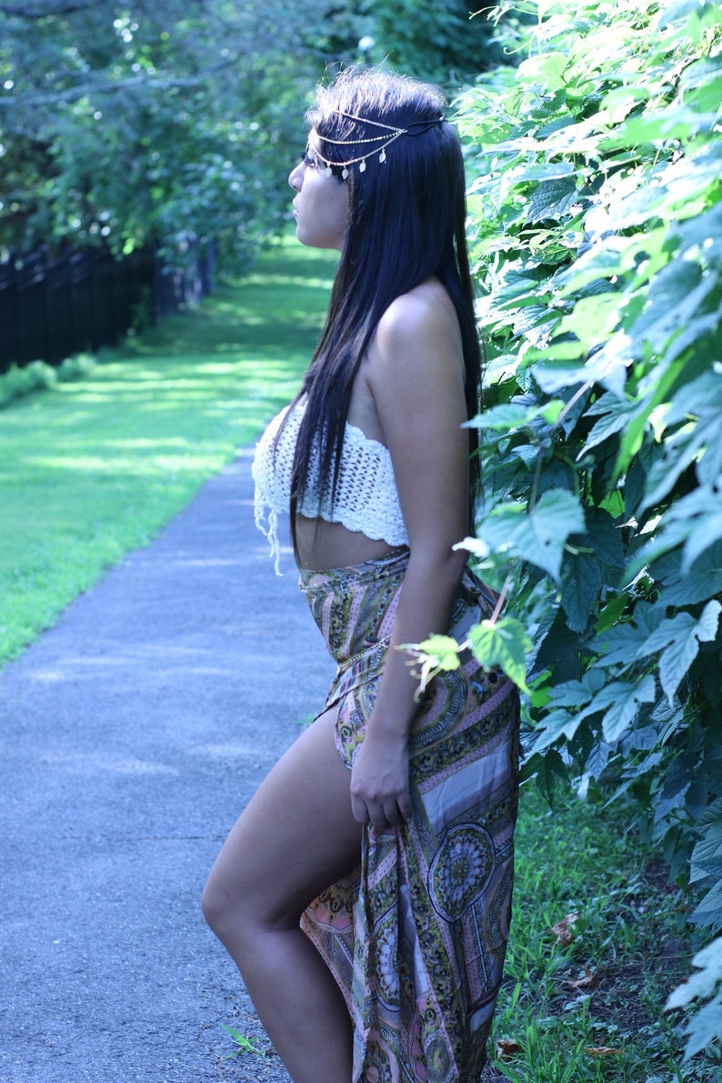 May include: A woman wearing a white crocheted top and a long, patterned skirt. She is standing in a forest setting with green foliage behind her. She is wearing a gold headpiece with small, dangling charms.