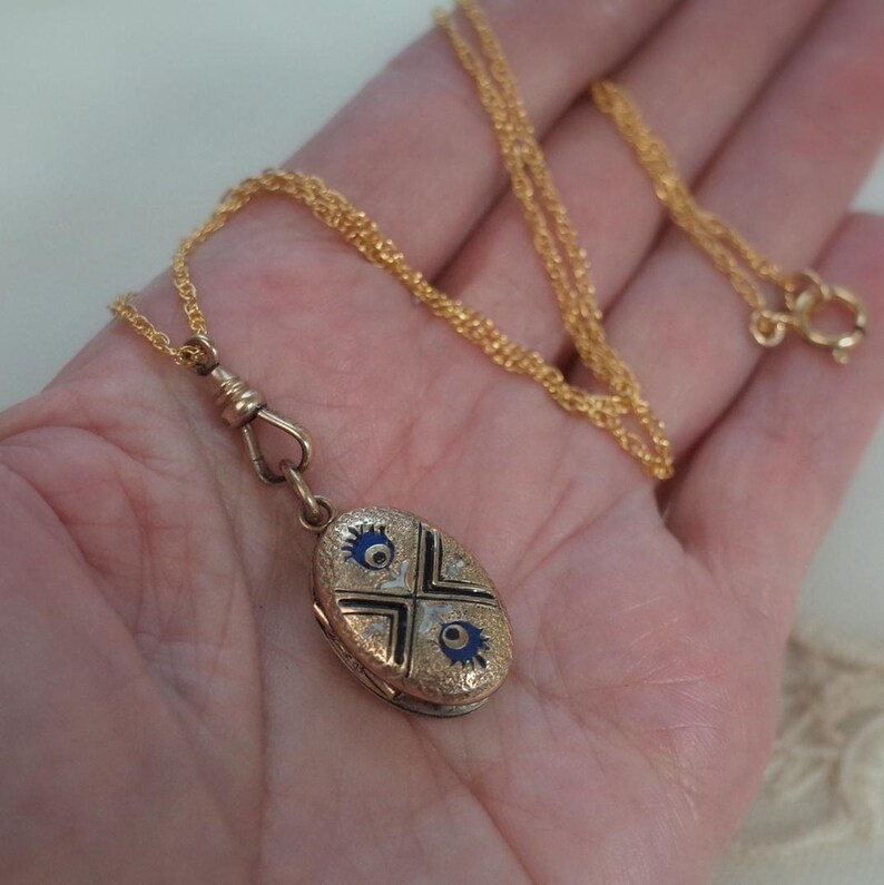 May include: Gold-toned oval pendant necklace with a delicate chain. The pendant features a cross design with blue and black enamel details. The necklace is held in a hand.