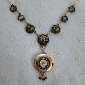 Puede incluir: Un collar dorado con un reloj colgante. El collar presenta colgantes circulares negros y dorados con dise&ntilde;os de paisajes. El colgante del reloj tiene un cristal central y peque&ntilde;as cuentas negras. El collar est&aacute; sobre una superficie de encaje blanco.