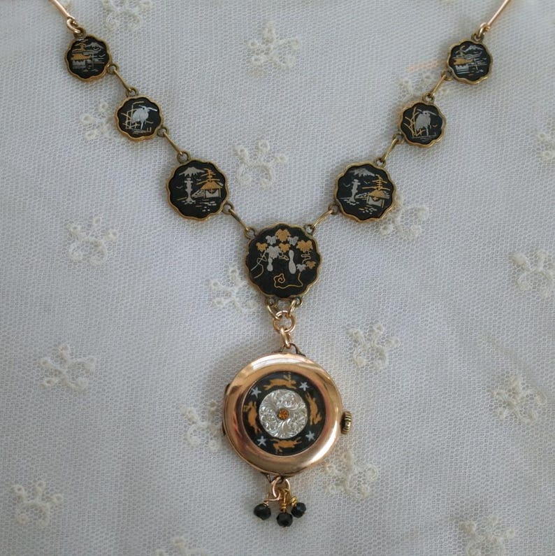 Puede incluir: Un collar dorado con un reloj colgante. El collar presenta colgantes circulares negros y dorados con dise&ntilde;os de paisajes. El colgante del reloj tiene un cristal central y peque&ntilde;as cuentas negras. El collar est&aacute; sobre una superficie de encaje blanco.