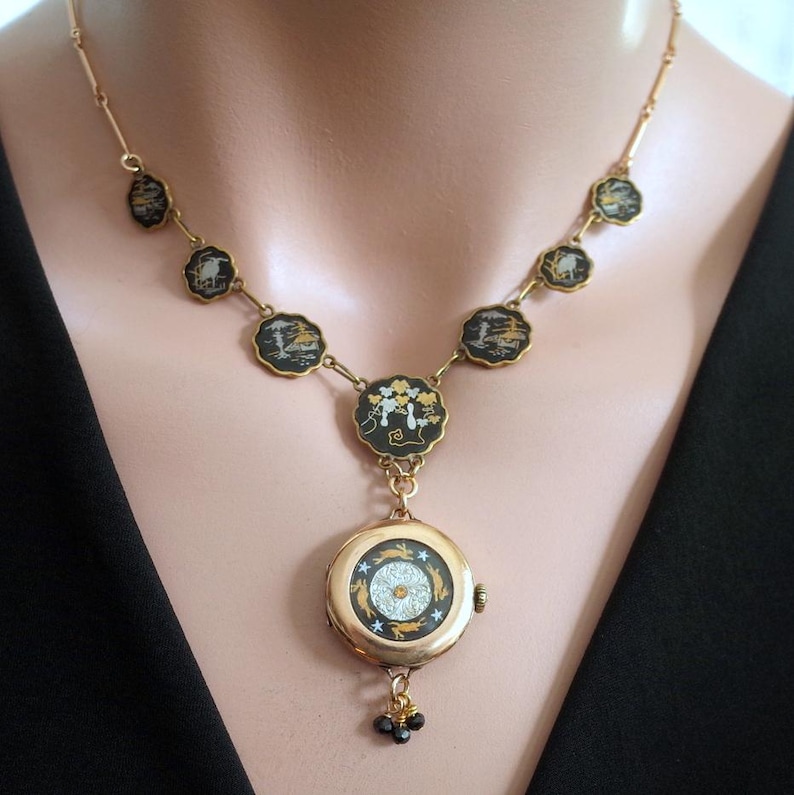 Puede incluir: Un collar dorado con un reloj colgante. El collar presenta colgantes circulares negros y dorados con dise&ntilde;os de paisajes. El reloj colgante tiene una esfera blanca y dorada con un dise&ntilde;o de estrella y un peque&ntilde;o grupo de cuentas negras.