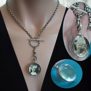 May include: A silver-toned necklace with a toggle clasp and a round pendant. The pendant contains a black and white photograph of a child. The necklace is worn against a black top.