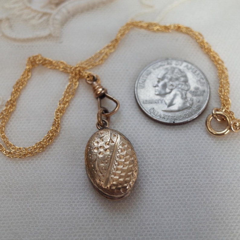 May include: A gold-colored oval locket pendant with intricate embossed detailing, suspended from a delicate gold chain. The locket is shown next to a U.S. quarter for size comparison. The chain has a clasp closure.