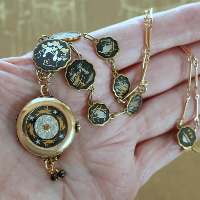Puede incluir: Un conjunto de reloj de bolsillo y collar dorados. La esfera del reloj presenta un dise&ntilde;o detallado con una gema naranja central. El collar tiene eslabones negros y dorados con temas florales y paisaj&iacute;sticos. El conjunto se sostiene en una mano.