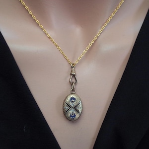 May include: A gold-toned necklace with a decorative oval pendant. The pendant features a detailed design with blue and white accents. The necklace is displayed against a black background.