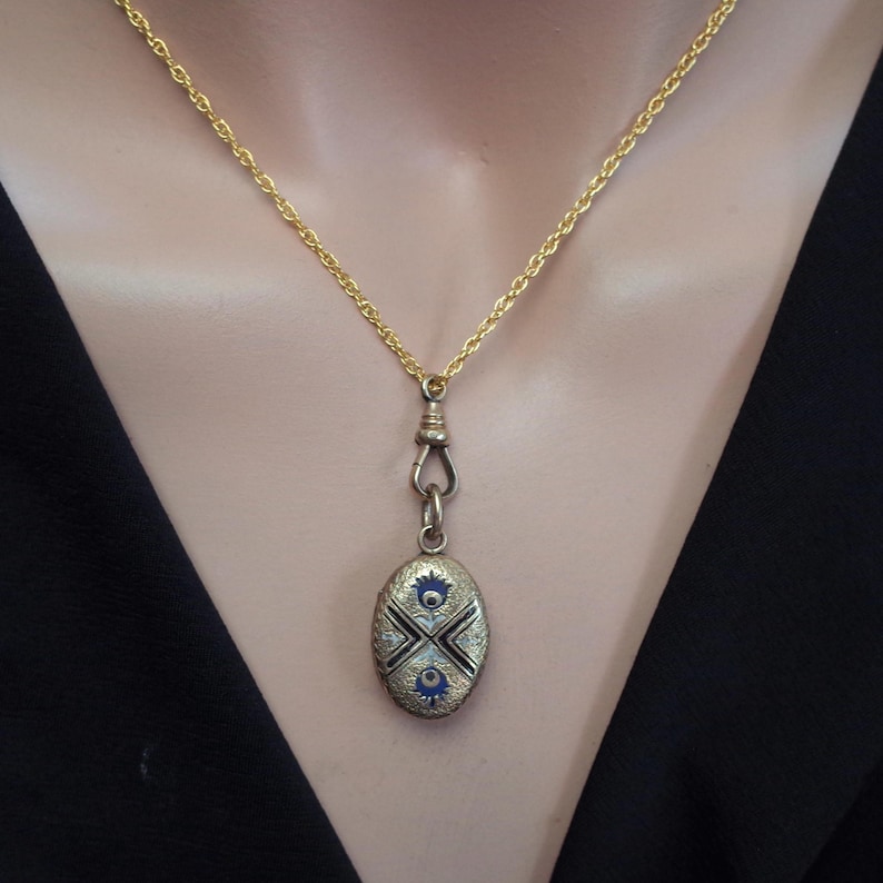 May include: A gold-toned necklace with a decorative oval pendant. The pendant features a detailed design with blue and white accents. The necklace is displayed against a black background.