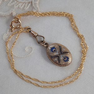 May include: Gold-toned necklace featuring an oval pendant with blue enamel accents and a cross design. The pendant is attached to a delicate chain with a lobster clasp. The necklace is displayed on a white, textured surface.