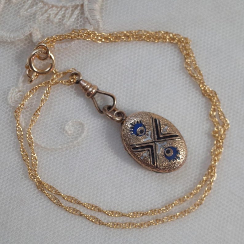 May include: Gold-toned necklace featuring an oval pendant with blue enamel accents and a cross design. The pendant is attached to a delicate chain with a lobster clasp. The necklace is displayed on a white, textured surface.