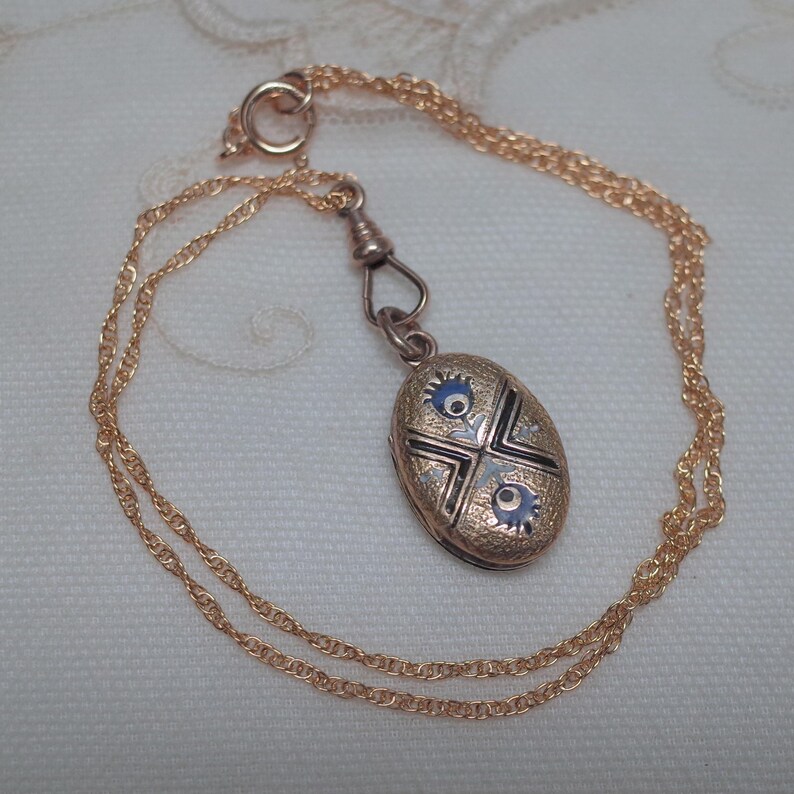 May include: A gold-toned oval locket pendant with blue and black enamel detailing, suspended from a delicate gold chain. The locket features a cross-like design with floral accents. The chain is approximately 20 inches long.
