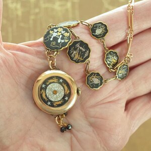 Puede incluir: Un reloj vintage dorado con una cadena decorativa. La esfera del reloj presenta un dise&ntilde;o central con una peque&ntilde;a gema naranja, rodeada por un borde negro con conejos y estrellas. La cadena tiene eslabones negros festoneados con dise&ntilde;os de paisajes.