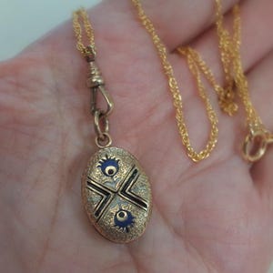 May include: Gold-toned necklace with an oval pendant. The pendant features a detailed design with black, white, and blue accents. The necklace has a delicate chain and a clasp. The pendant is approximately 1 inch in length.