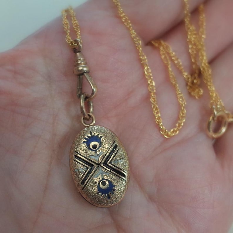 May include: Gold-toned necklace with an oval pendant. The pendant features a detailed design with black, white, and blue accents. The necklace has a delicate chain and a clasp. The pendant is approximately 1 inch in length.