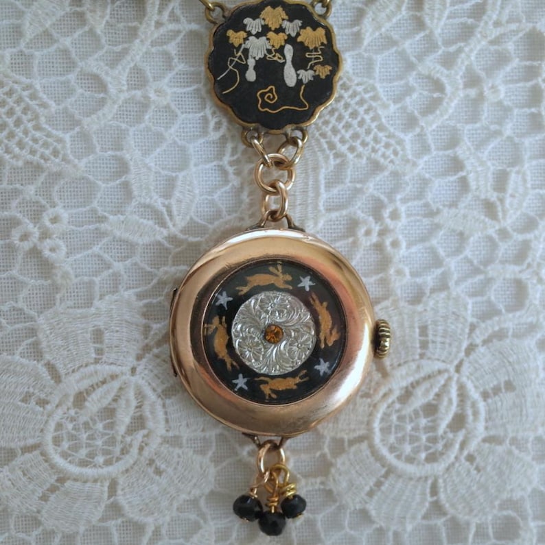 Puede incluir: Un collar con colgante dorado con un colgante redondo tipo reloj y una pieza superior circular m&aacute;s peque&ntilde;a. El colgante presenta un fondo negro con conejos y estrellas doradas, y un centro plateado y naranja. La pieza superior tiene un fondo negro con dise&ntilde;os florales dorados y blancos.