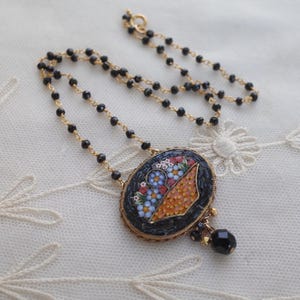 May include: A gold-toned necklace with a black beaded chain and a mosaic pendant. The pendant features a floral design in shades of blue, orange, and white, set against a black background. A black teardrop-shaped bead hangs below the pendant.