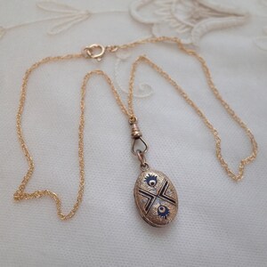 May include: A gold-toned necklace with a detailed oval pendant. The pendant features a blue and black design with intricate detailing. The chain is delicate and the clasp is visible. The necklace is displayed on a white, textured surface.