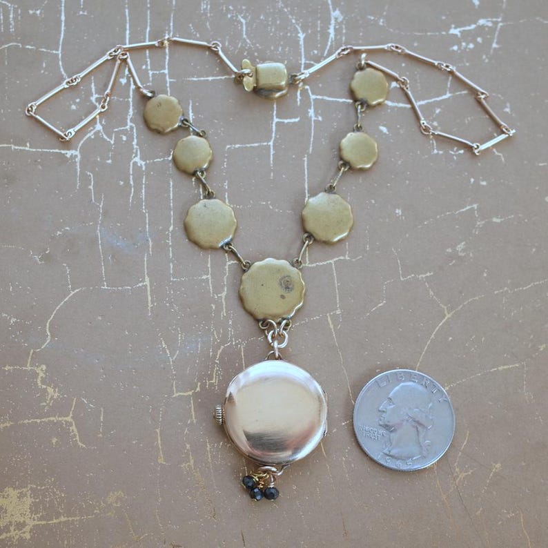 Puede incluir: Un collar vintage dorado con un colgante de medall&oacute;n redondo y una serie de amuletos circulares con bordes festoneados. El collar incluye un peque&ntilde;o grupo de cuentas negras y una moneda para comparar el tama&ntilde;o. La cadena tiene un dise&ntilde;o de eslabones rectangulares.