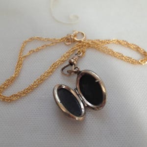 May include: A gold-colored locket necklace with an open oval pendant. The pendant reveals a dark interior. The chain is a delicate, twisted design. The locket is open, revealing the dark interior. The necklace is on a white surface.