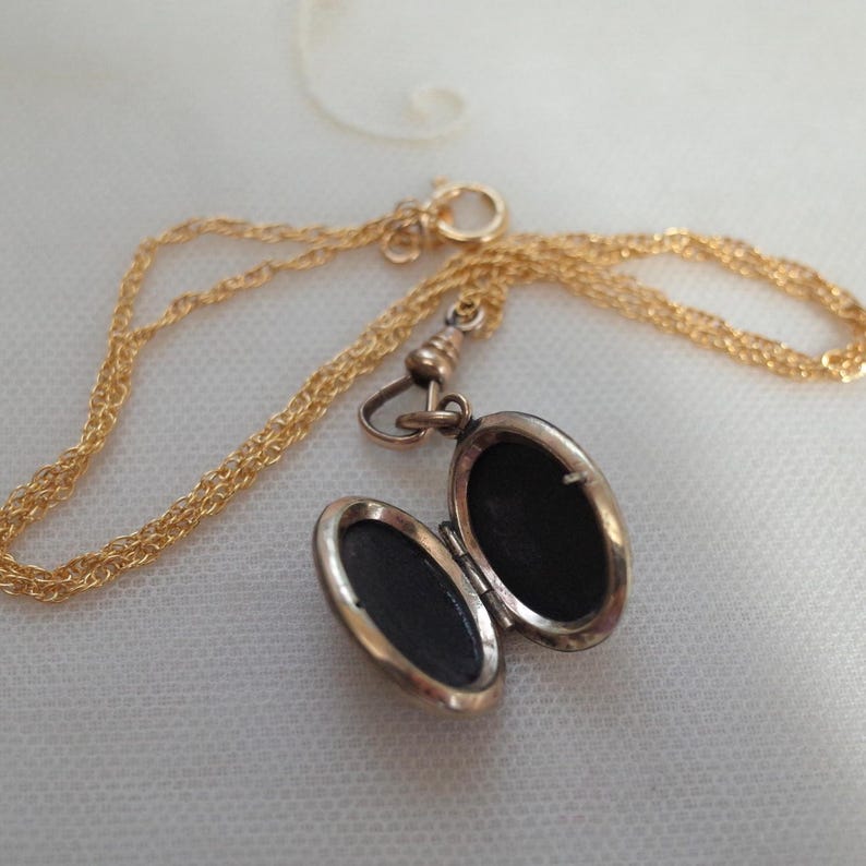 May include: A gold-colored locket necklace with an open oval pendant. The pendant reveals a dark interior. The chain is a delicate, twisted design. The locket is open, revealing the dark interior. The necklace is on a white surface.