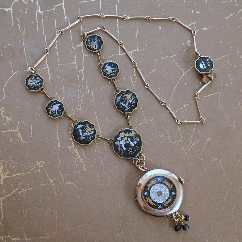 Puede incluir: Un collar dorado con colgantes negros festoneados con dise&ntilde;os de paisajes en blanco y dorado. El collar incluye un colgante redondo con dise&ntilde;o de esfera de reloj y peque&ntilde;os detalles de cuentas negras.