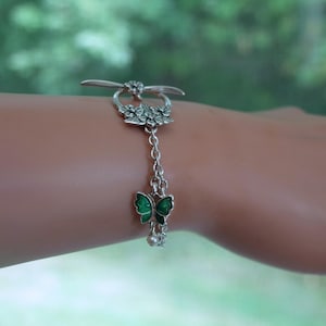 May include: A silver-toned toggle bracelet featuring a floral-shaped clasp and a small green enamel butterfly charm. The bracelet is constructed with a delicate metal chain and includes a tiny white bead accent dangling near the butterfly charm.