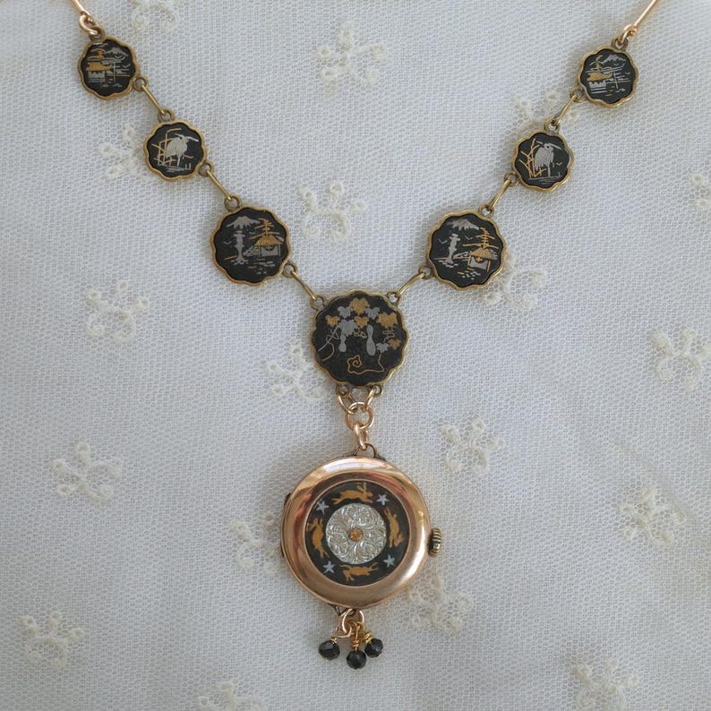 Puede incluir: Un collar dorado con colgantes circulares negros y dorados con dise&ntilde;os de paisajes. El collar tiene un colgante redondo con un dise&ntilde;o de esfera de reloj y peque&ntilde;os colgantes de cuentas negras.