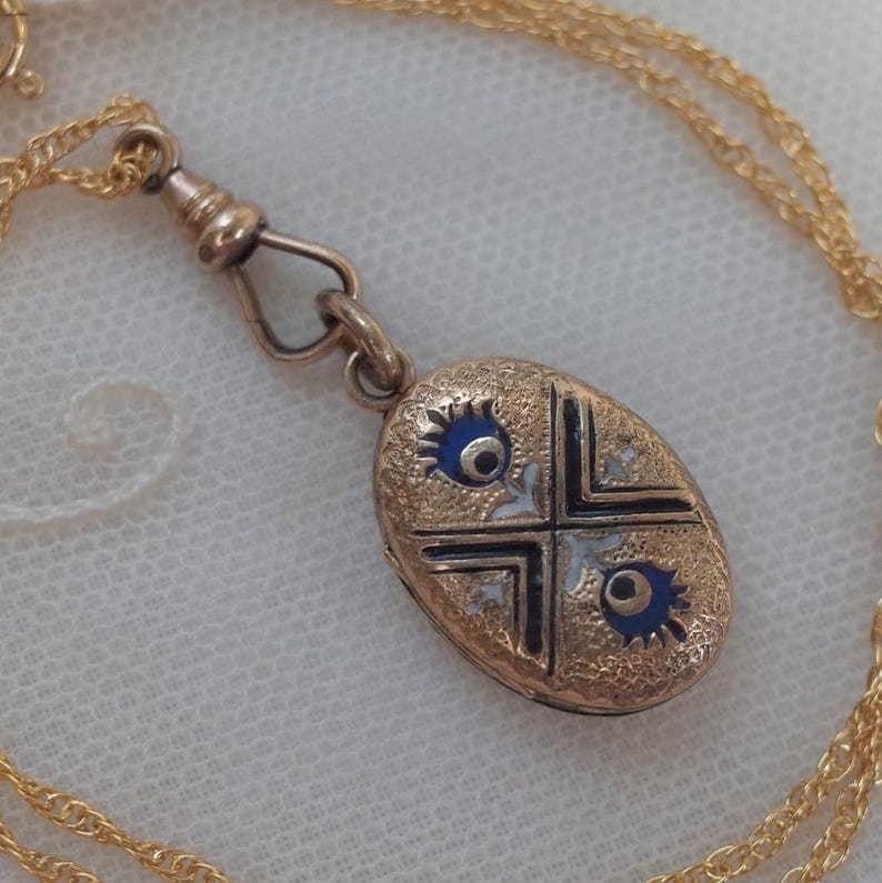 May include: Gold-toned oval pendant necklace with a decorative design. The pendant features a cross design with blue accents and intricate detailing. The necklace has a delicate chain and a clasp.