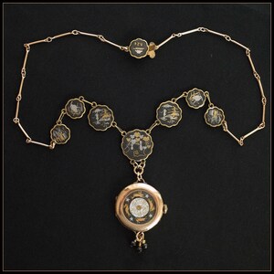 Puede incluir: Un collar dorado con un reloj colgante. El collar presenta una serie de amuletos circulares negros y dorados con dise&ntilde;os de paisajes. El reloj colgante tiene una esfera negra con detalles dorados y plateados y un peque&ntilde;o grupo de cuentas negras.