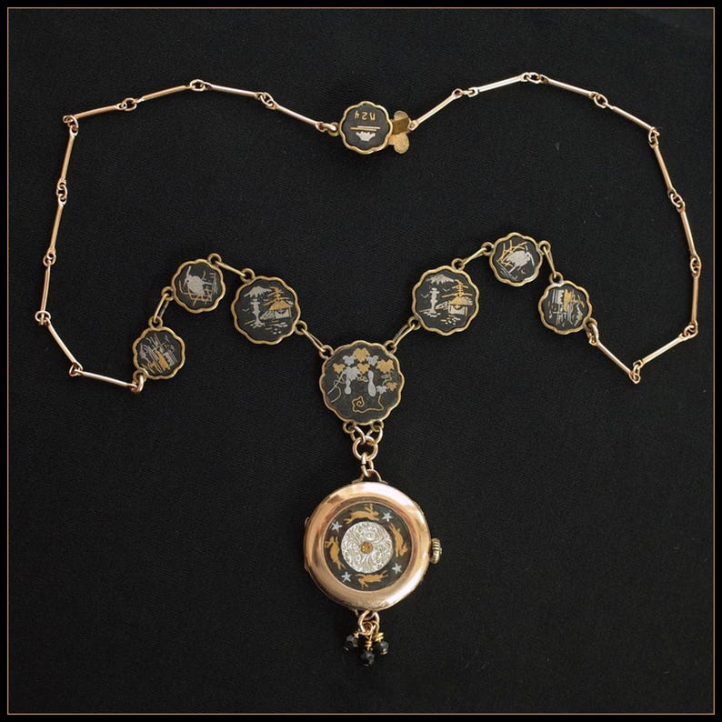 Puede incluir: Un collar dorado con un reloj colgante. El collar presenta una serie de amuletos circulares negros y dorados con dise&ntilde;os de paisajes. El reloj colgante tiene una esfera negra con detalles dorados y plateados y un peque&ntilde;o grupo de cuentas negras.