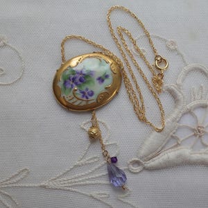 May include: A gold-toned necklace featuring an oval pendant with a hand-painted floral design in purple and green. The pendant is suspended from a delicate chain, with a dangling charm of a gold bead, purple beads, and a teardrop-shaped crystal.