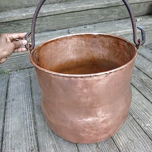May include: A copper cauldron with a dark metal handle. The cauldron has a rounded body and a wide opening. The handle is attached to the sides of the cauldron with metal loops. The cauldron is sitting on a wooden surface.